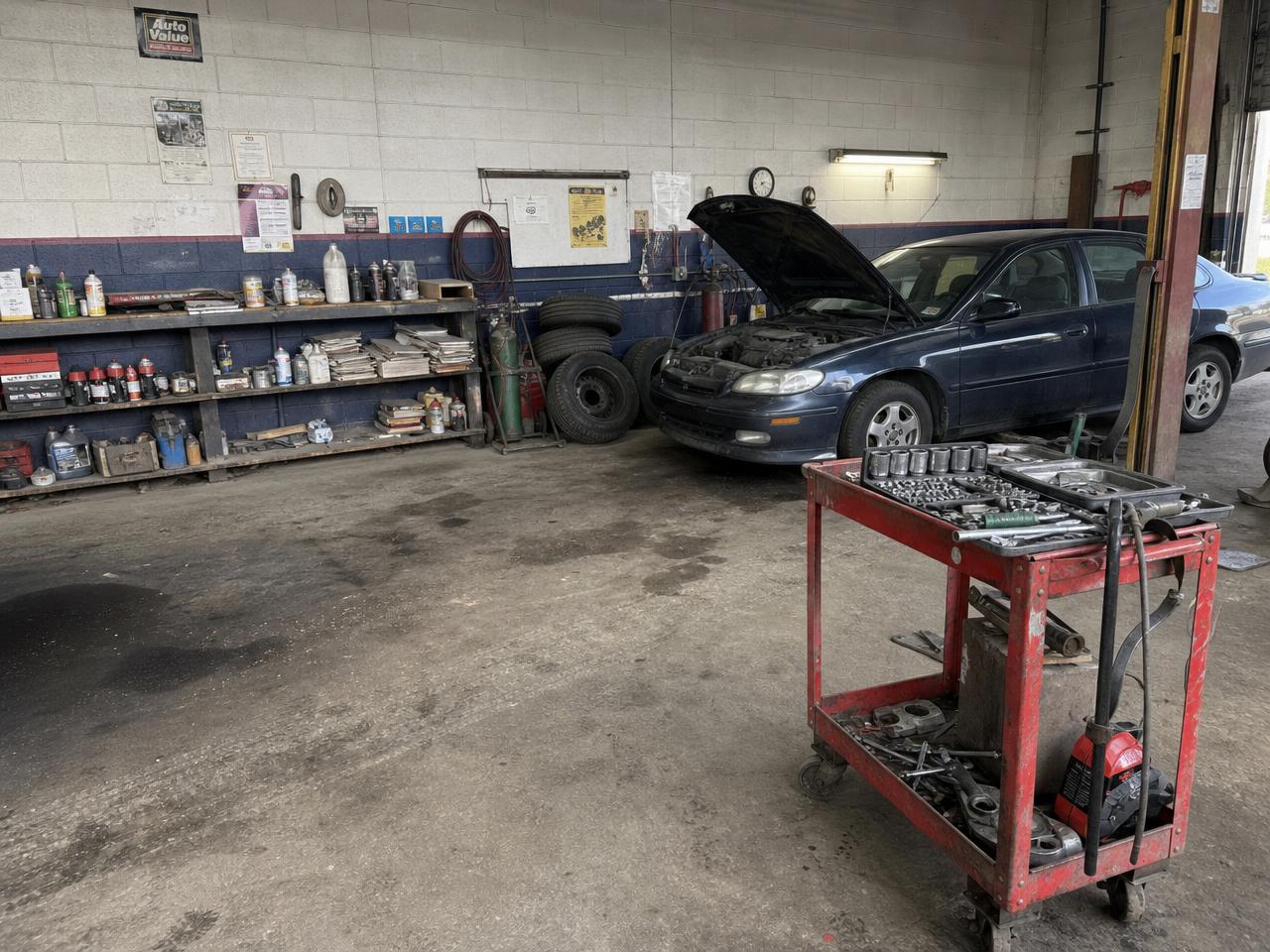 A typical uncertified repair bay: stained concrete floor, cluttered shelves, tools left out, and an older vehicle with its hood up